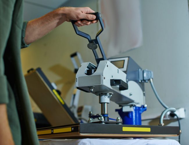Cropped view of young craftsman using screen printing machine on clothes while working in blurred print studio at background, customer-focused small business concept